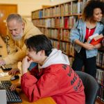 Estudantes colaboram diante de computadores em uma biblioteca, entre livros e mesas de estudo.