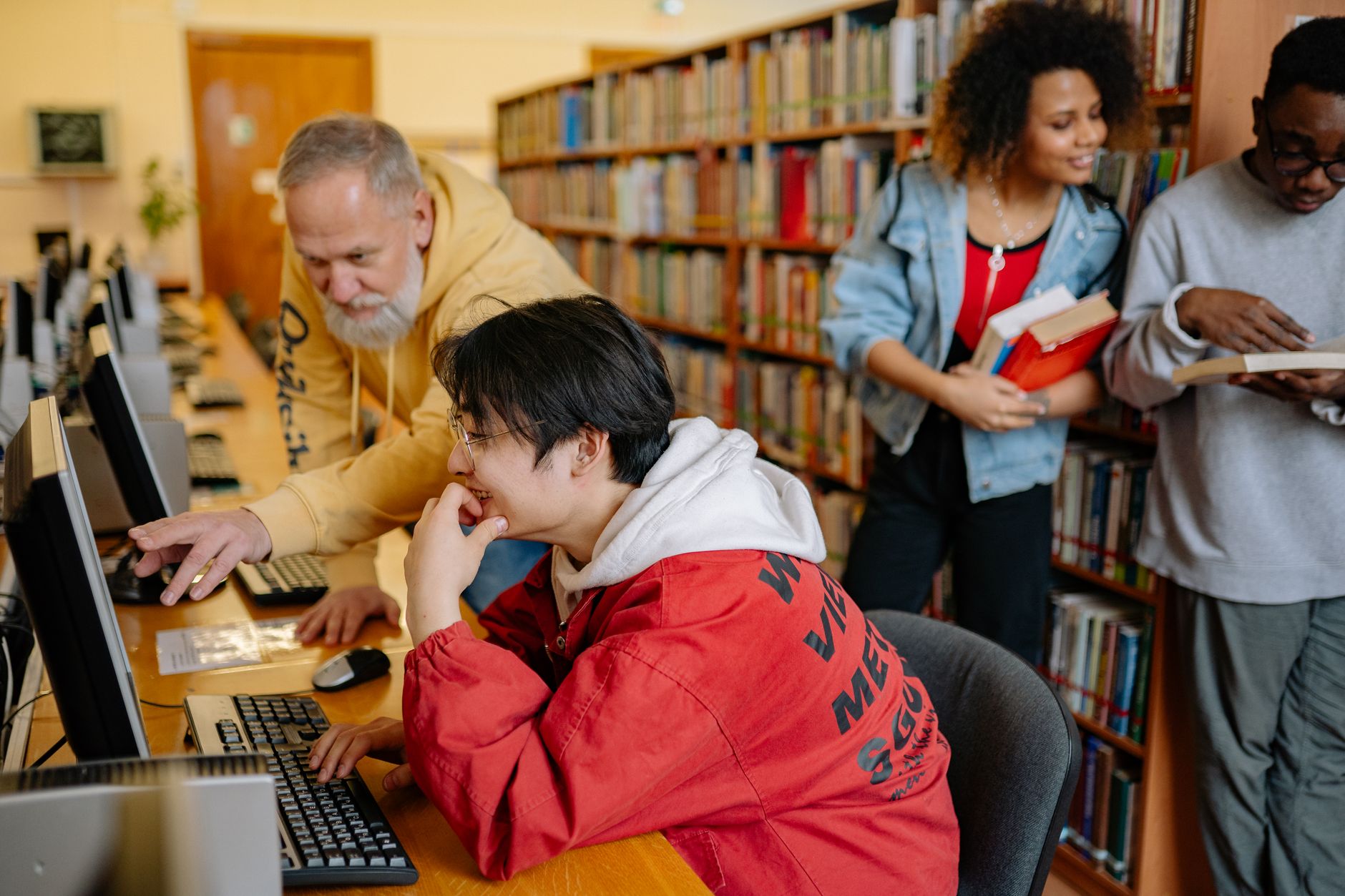 Estudantes colaboram diante de computadores em uma biblioteca, entre livros e mesas de estudo.