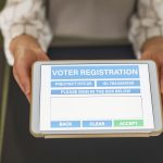 Mãos seguram um tablet que exibe um formulário digital de registro eleitoral.