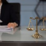 Balança da Justiça sobre uma mesa de escritório, com uma profissional desfocada ao fundo