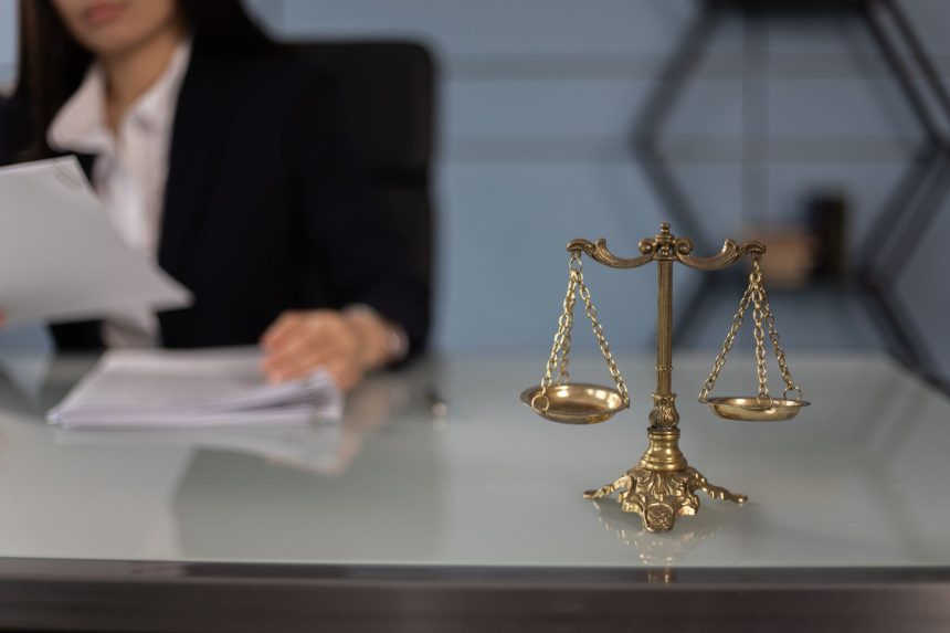 Balança da Justiça sobre uma mesa de escritório, com uma profissional desfocada ao fundo