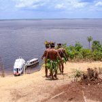 Dois homens indígenas usando trajes tradicionais observam barcos no rio Amazonas.