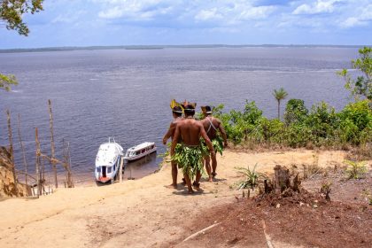 Dois homens indígenas usando trajes tradicionais observam barcos no rio Amazonas.