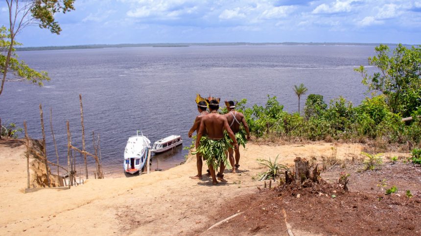 Dois homens indígenas usando trajes tradicionais observam barcos no rio Amazonas.