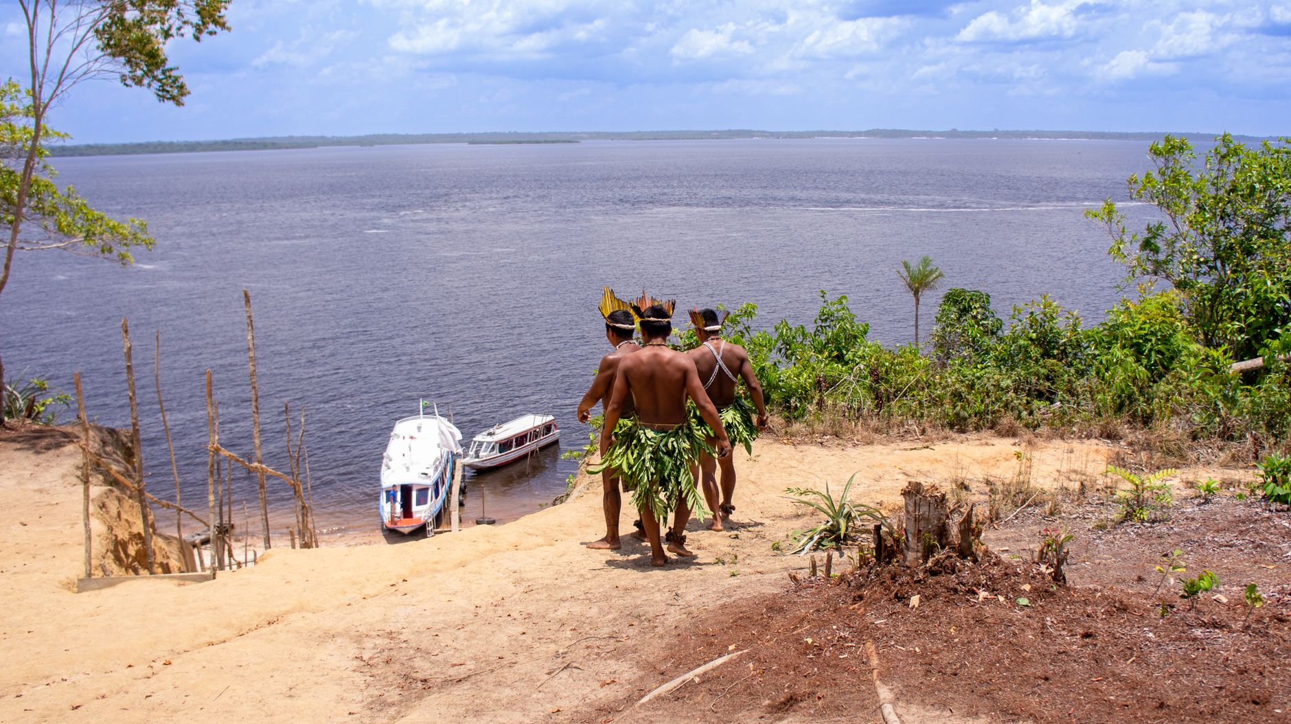 Dois homens indígenas usando trajes tradicionais observam barcos no rio Amazonas.