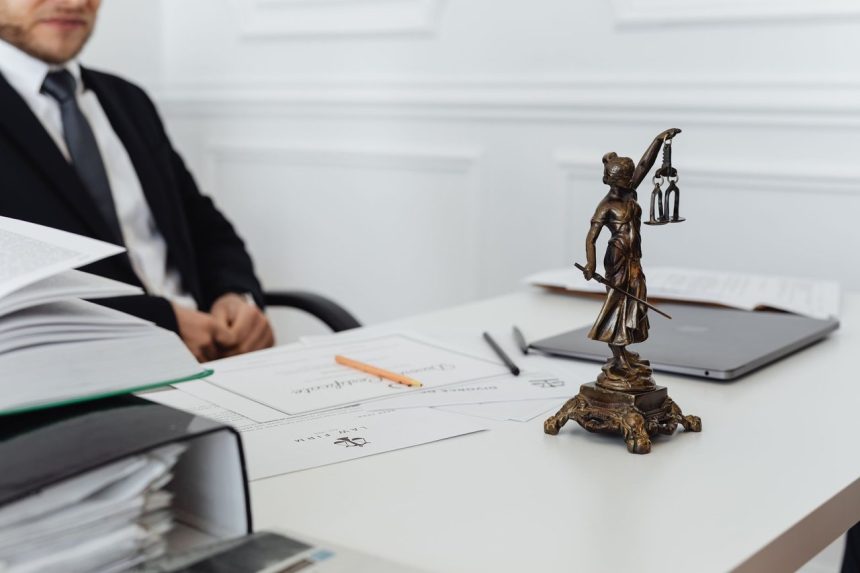 Mesa de trabalho jurídico com documentos, laptop e uma estátua da Justiça ao fundo