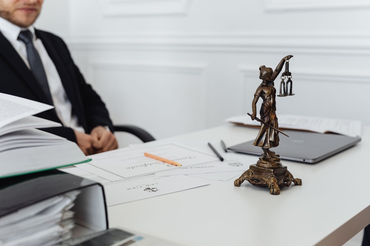 Mesa de trabalho jurídico com documentos, laptop e uma estátua da Justiça ao fundo