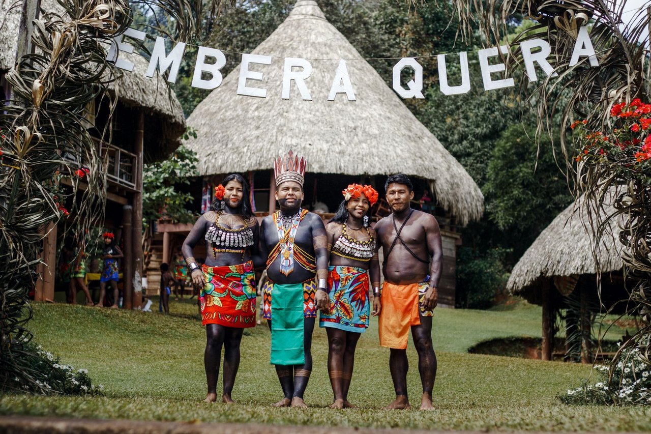 Grupo de pessoas indígenas Emberá posa em trajes tradicionais diante de sua comunidade.