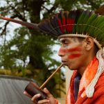 Pessoa indígena usando cocar colorido e fumando um cachimbo em São Paulo.