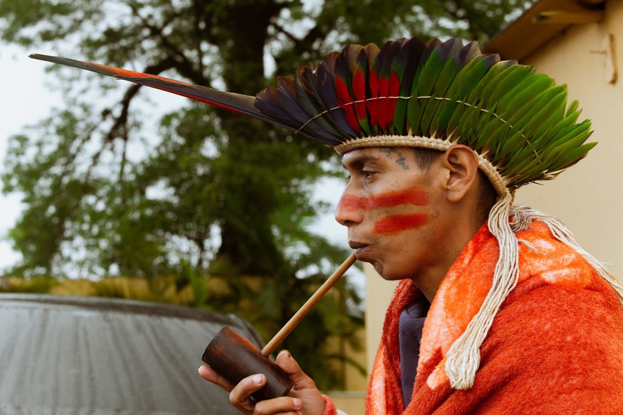 Pessoa indígena usando cocar colorido e fumando um cachimbo em São Paulo.
