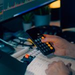 Mesa de trabalho com calculadora e monitor exibindo gráficos financeiros, representando a análise de dados e a conferência de informações empresariais.