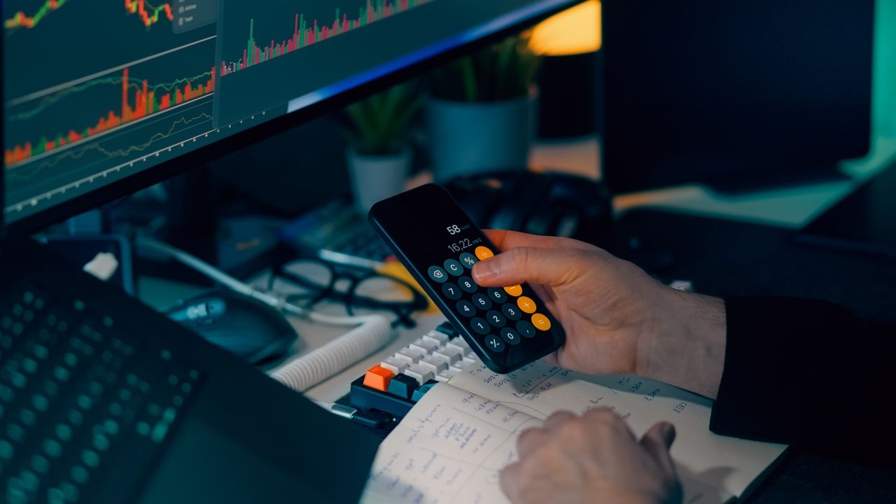 Mesa de trabalho com calculadora e monitor exibindo gráficos financeiros, representando a análise de dados e a conferência de informações empresariais.
